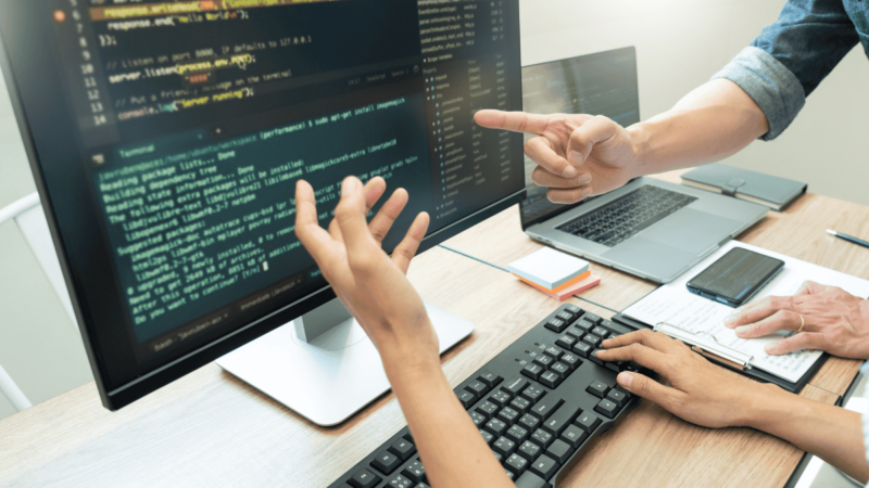 Two people collaborating on coding in a workspace, with one person gesturing toward a large monitor displaying lines of code. A laptop, notepad, smartphone, and other tools are visible on the desk, creating a productive and focused environment for learning and development.